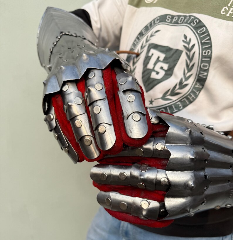 K&ouml;nnte beinhalten: Ein Paar gegliederte Stahlhandschuhe mit rotem Stofffutter. Die Handschuhe bestehen aus genieteten Metallplatten und dienen zum Schutz der H&auml;nde und Unterarme. Das Metall hat eine polierte, reflektierende Oberfl&auml;che.