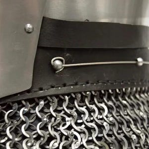 May include: Close-up of a medieval helmet with a metal plate, black leather trim, and chainmail. The helmet features a riveted construction and a silver wire detail. The chainmail is made of interlocking metal rings.