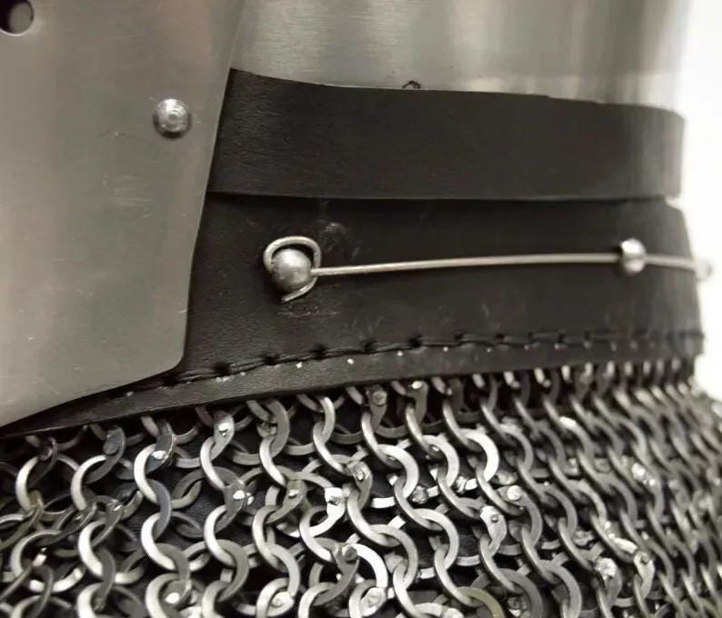 May include: Close-up of a medieval helmet with a metal plate, black leather trim, and chainmail. The helmet features a riveted construction and a silver wire detail. The chainmail is made of interlocking metal rings.