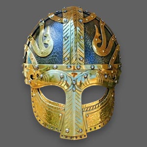 May include: A gold and silver Viking helmet with intricate details and a face mask. The helmet is decorated with a pattern of stylized animal heads and geometric shapes.