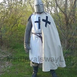 May include: A full-body shot of a medieval knight in armor. The knight is wearing a metal helmet, chainmail, and a white tunic with a black cross. A white cape with a black cross is draped over the shoulders. The knight is standing in a grassy area with trees in the background.