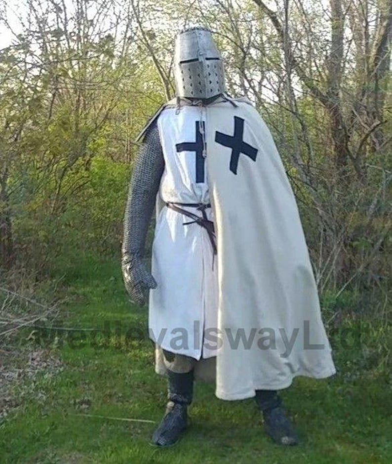 May include: A full-body shot of a medieval knight in armor. The knight is wearing a metal helmet, chainmail, and a white tunic with a black cross. A white cape with a black cross is draped over the shoulders. The knight is standing in a grassy area with trees in the background.