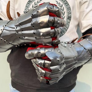 K&ouml;nnte beinhalten: Ein Paar gegliederte Stahlhandschuhe mit poliertem Finish. Die Innenseite ist mit rotem Stoff gef&uuml;ttert. Die Handschuhe sind mit mehreren Platten und Nieten versehen und dienen wahrscheinlich der historischen Nachstellung oder Ausstellung.