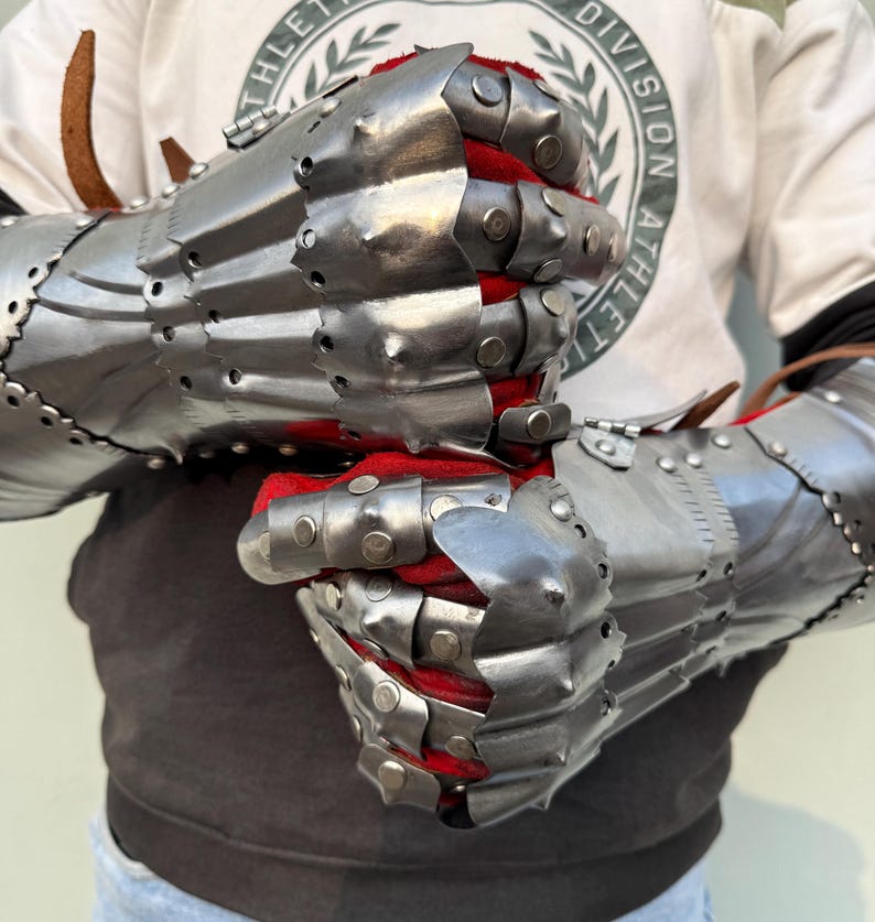 K&ouml;nnte beinhalten: Ein Paar gegliederte Stahlhandschuhe mit poliertem Finish. Die Innenseite ist mit rotem Stoff gef&uuml;ttert. Die Handschuhe sind mit mehreren Platten und Nieten versehen und dienen wahrscheinlich der historischen Nachstellung oder Ausstellung.