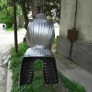May include: A silver metal suit of armor with a black leather skirt. The armor is hanging from a hook on a wall.