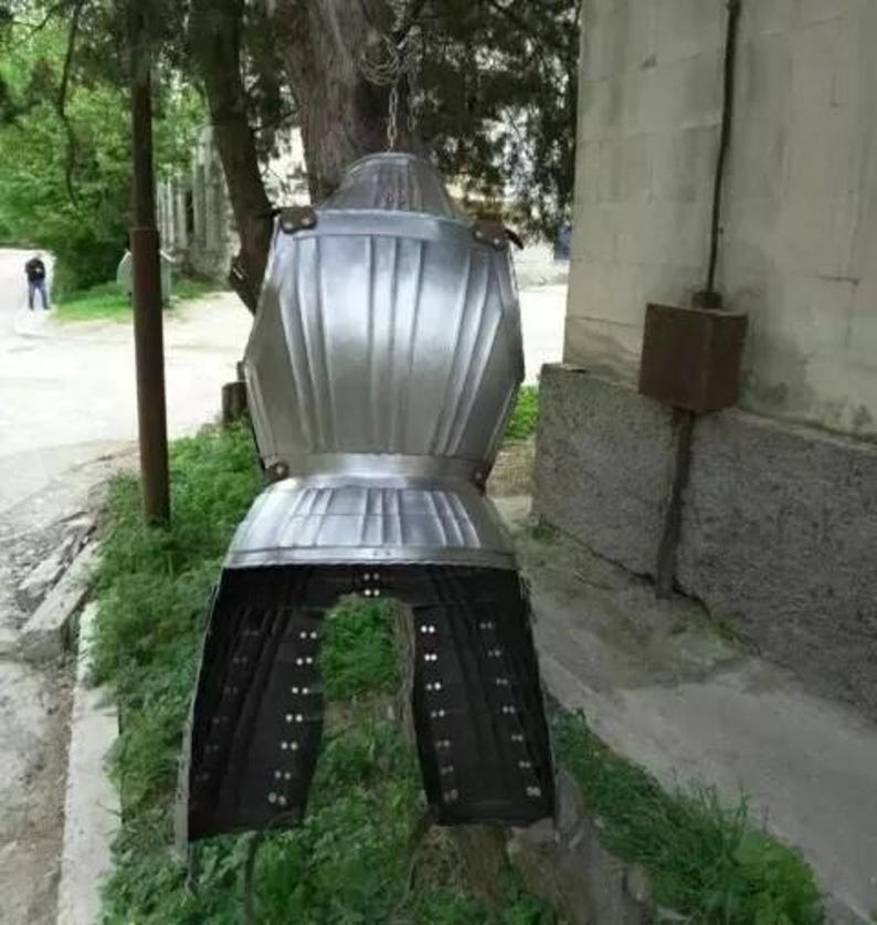 May include: A silver metal suit of armor with a black leather skirt. The armor is hanging from a hook on a wall.