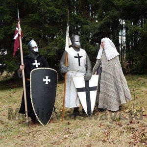 May include: Three people in medieval costumes stand in a forest. Two are dressed as knights, wearing helmets, chainmail, and tunics with crosses. They hold shields and spears. The third person wears a long, gray cloak and a white head covering.
