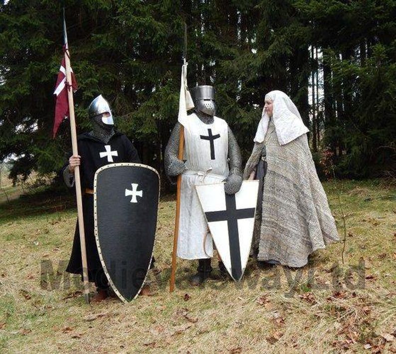 May include: Three people in medieval costumes stand in a forest. Two are dressed as knights, wearing helmets, chainmail, and tunics with crosses. They hold shields and spears. The third person wears a long, gray cloak and a white head covering.