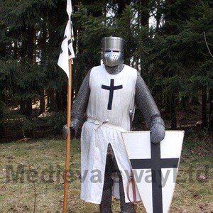 May include: A medieval knight in full armor stands holding a spear with a flag and a shield. The knight wears a metal helmet, chainmail, and a white tunic with a black cross. The shield is white with a black cross. The scene is set outdoors.