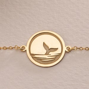 Può includere: Un braccialetto a catena dorata con un ciondolo circolare che presenta una silhouette di coda di balena.