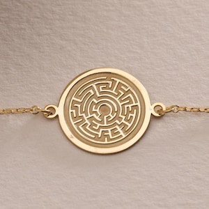 Puede incluir: Un brazalete de cadena dorada con un colgante circular que presenta un diseño de laberinto detallado.