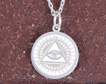 Puede incluir: Collar con colgante plateado. El colgante circular presenta un diseño de rayos de sol y un triángulo con un ojo en el centro. La cadena es fina y el conjunto se encuentra sobre un fondo texturizado marrón rojizo.