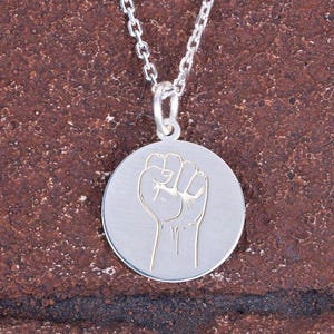 May include: Silver-toned pendant necklace featuring a raised fist design. The pendant is circular with a detailed fist illustration in a contrasting color. The chain is delicate and silver, suitable for everyday wear.