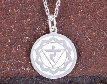 May include: Silver-colored pendant necklace featuring a circular charm with a detailed, symmetrical design. The charm has a central geometric shape surrounded by a floral-like border. The necklace chain is also silver and hangs against a textured, reddish-brown background.
