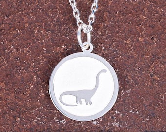 May include: A silver necklace featuring a round pendant with a dinosaur silhouette. The pendant has a white background and a silver outline. The chain is also silver and hangs against a textured brown background.