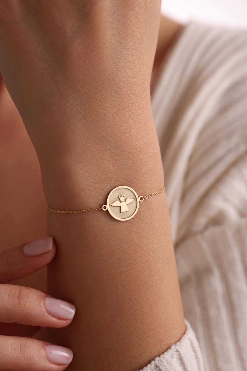 K&ouml;nnte beinhalten: Ein goldenes Kettenarmband mit einem runden Anh&auml;nger, der eine Engelsilhouette zeigt.