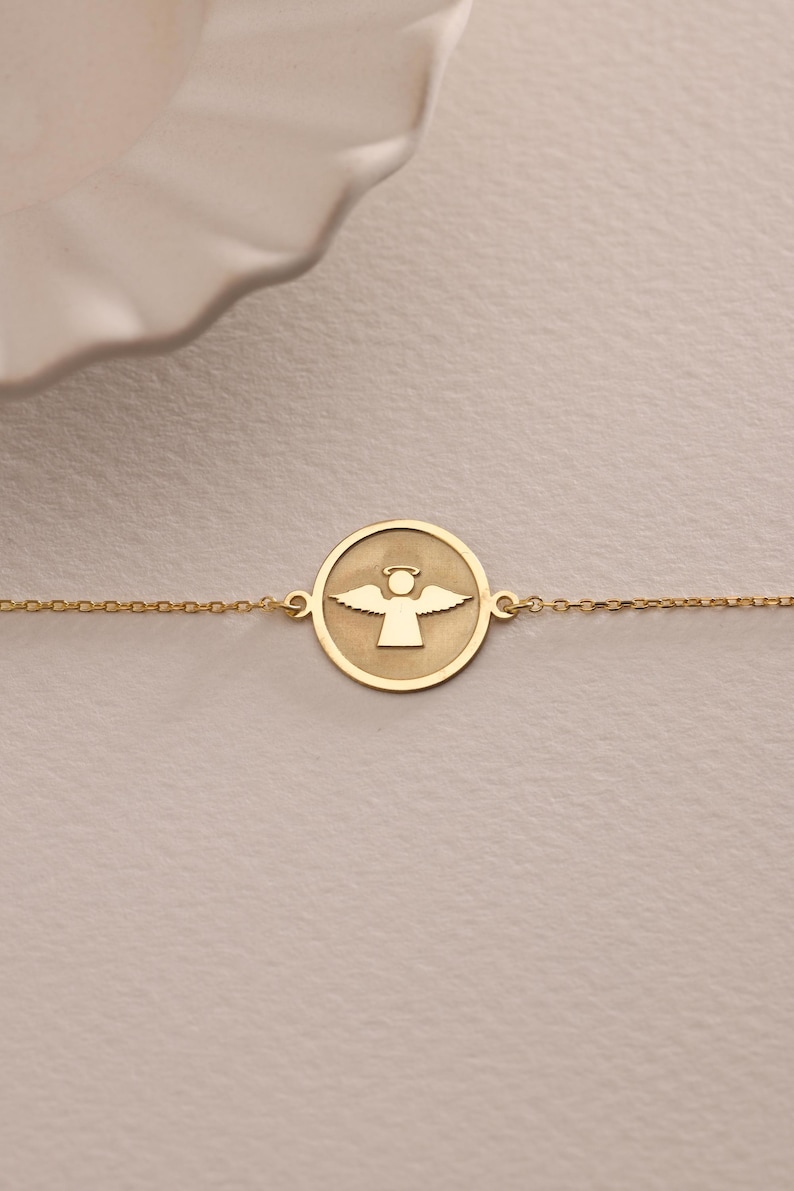 K&ouml;nnte beinhalten: Ein goldenes Kettenarmband mit einem runden Anh&auml;nger, der ein ausgeschnittenes Design eines Engels mit Fl&uuml;geln zeigt.