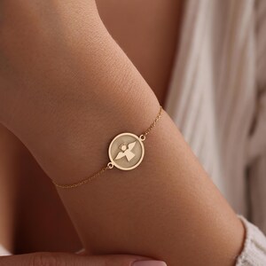 K&ouml;nnte beinhalten: Ein goldenes Kettenarmband mit einem runden Anh&auml;nger, der ein ausgeschnittenes Engelsmotiv zeigt.