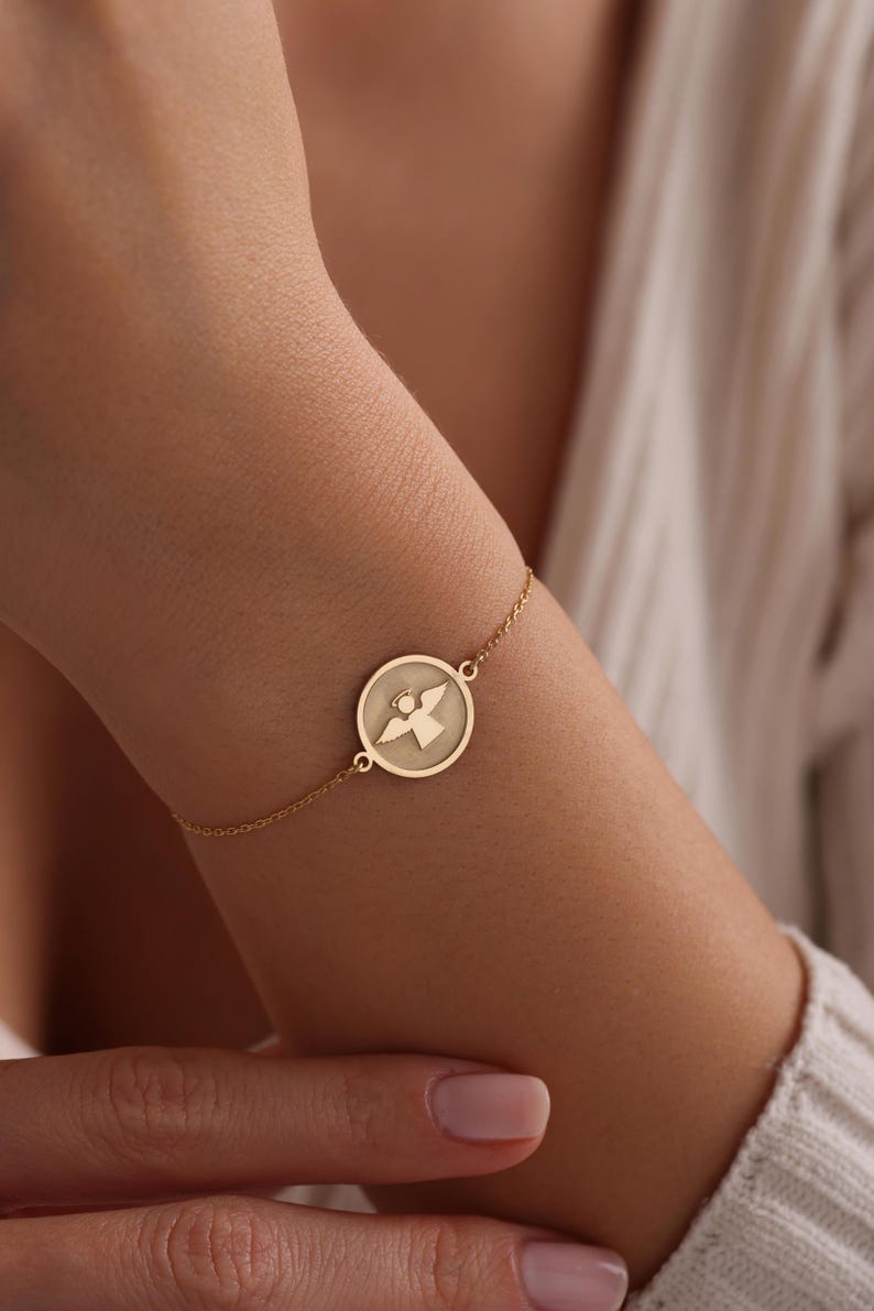 K&ouml;nnte beinhalten: Ein goldenes Kettenarmband mit einem runden Anh&auml;nger, der ein ausgeschnittenes Engelsmotiv zeigt.