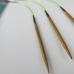May include: Close-up of four bamboo circular knitting needles with green flexible cords. The needles have a natural wood finish and are connected to the cords with black connectors. The needles are labeled with text.