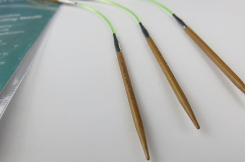 May include: Close-up of four bamboo circular knitting needles with green flexible cords. The needles have a natural wood finish and are connected to the cords with black connectors. The needles are labeled with text.
