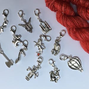 May include: A selection of silver charms with lobster claw clasps. The charms feature a fairy, a castle, a lion, a monkey, an axe, a high-heeled shoe, a jester, a hot air balloon, and a girl with a basket. A skein of red yarn is in the background.