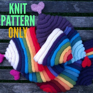 Puede incluir: Patrón de tejido para un gorro de ganchillo de colores arcoíris con un corazón en la parte superior. El gorro está hecho de varios colores de hilo, incluyendo rojo, naranja, amarillo, verde, azul, añil y violeta. El gorro está sobre una superficie de madera.