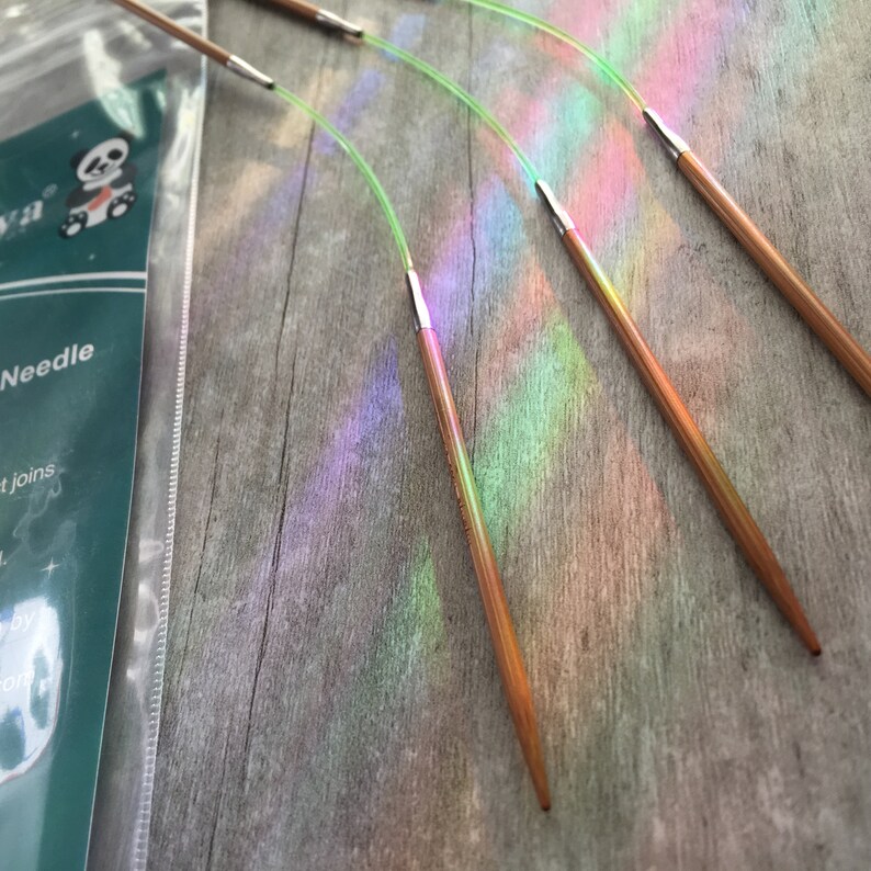 May include: Close-up of several wooden knitting needles with metal tips and flexible green cords. The needles are arranged diagonally across a gray wooden surface, with a package visible in the background. The needles are a light brown color.