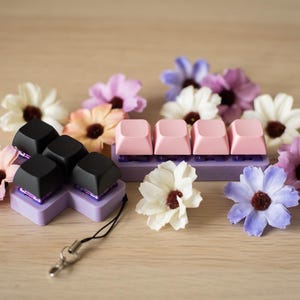 May include: A set of black and pink keycaps for a mechanical keyboard. The keycaps are arranged in a flower shape and are surrounded by artificial flowers.
