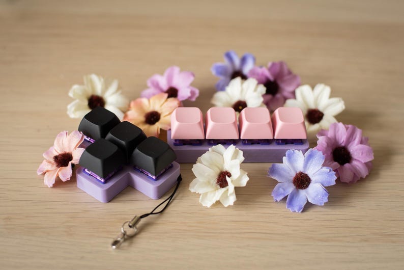 May include: A set of black and pink keycaps for a mechanical keyboard. The keycaps are arranged in a flower shape and are surrounded by artificial flowers.