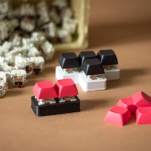 May include: A collection of mechanical keyboard keycaps in various colors, including black, white, and red. The keycaps are arranged in different configurations, with some stacked on top of each other. The keycaps are made of plastic and have a smooth, glossy finish.