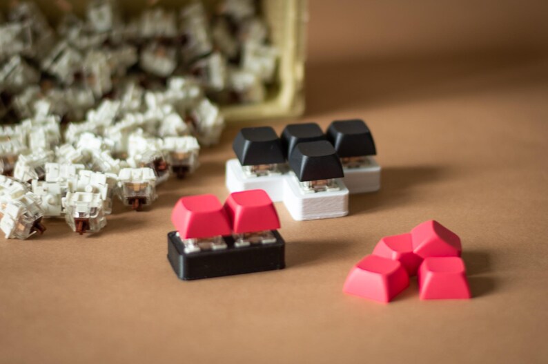 May include: A collection of mechanical keyboard keycaps in various colors, including black, white, and red. The keycaps are arranged in different configurations, with some stacked on top of each other. The keycaps are made of plastic and have a smooth, glossy finish.