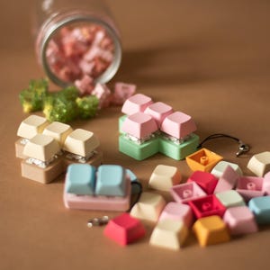 May include: Assortment of colorful keycaps in various shapes and sizes, including a set of pastel pink, green, and white keycaps, a set of light blue and pink keycaps, and a set of beige and white keycaps. The keycaps are arranged on a brown surface.