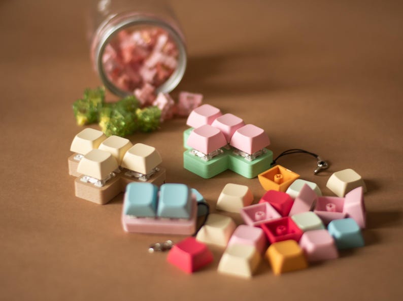 May include: Assortment of colorful keycaps in various shapes and sizes, including a set of pastel pink, green, and white keycaps, a set of light blue and pink keycaps, and a set of beige and white keycaps. The keycaps are arranged on a brown surface.
