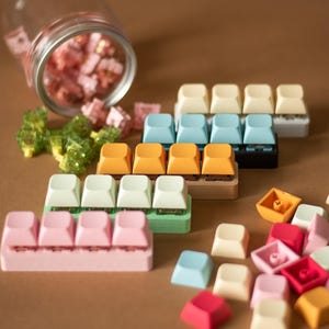 May include: Assortment of colorful keycaps for mechanical keyboards. The keycaps are arranged in rows and include various colors such as pink, green, blue, and orange. Some keycaps are in a jar, while others are scattered on a brown surface.