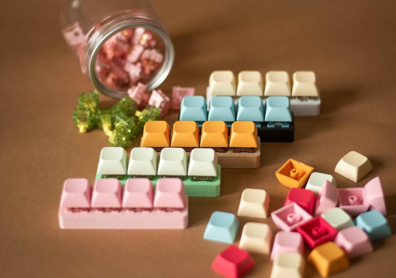 May include: Assortment of colorful keycaps for mechanical keyboards. The keycaps are arranged in rows and include various colors such as pink, green, blue, and orange. Some keycaps are in a jar, while others are scattered on a brown surface.