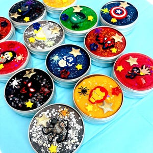 May include: A selection of round tins filled with colourful, glittery slime, each decorated with superhero-themed embellishments. The tins feature characters like Bat hero, American hero, and web crawler, with star and spider accents.