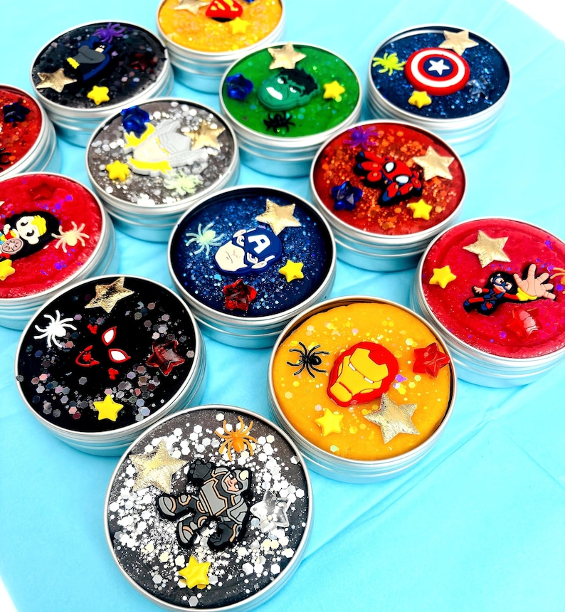 May include: A selection of round tins filled with colourful, glittery slime, each decorated with superhero-themed embellishments. The tins feature characters like Bat hero, American hero, and web crawler, with star and spider accents.