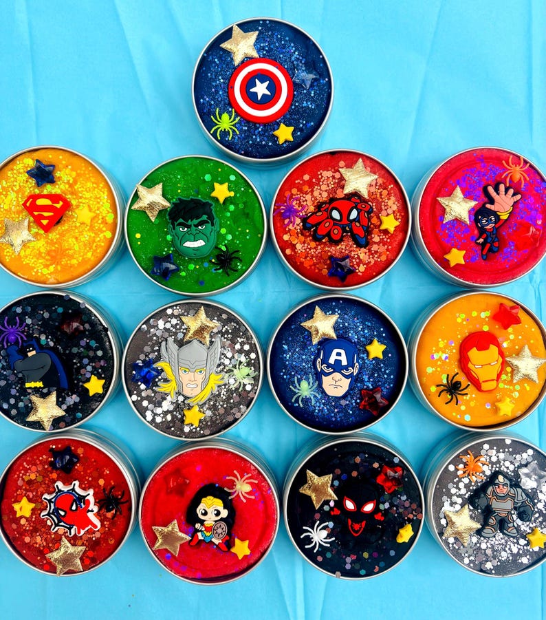 May include: A selection of round containers filled with glittery slime in various colours, each adorned with a superhero emblem and star-shaped decorations. The containers feature characters such as American superhero and Web-crawler.