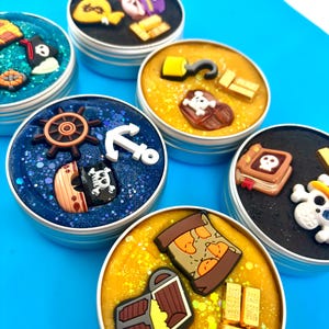 May include: Five round metal tins filled with colourful slime, each topped with various pirate-themed charms including gold bars, treasure chests, skulls, anchors, ship wheels, and maps. The slimes feature glittery textures in shades of blue, yellow, and black.