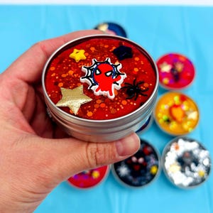 May include: A silver tin filled with red slime, decorated with a web-crawler emblem, gold and yellow stars, and a black spider. Other tins with colourful contents are in the background.