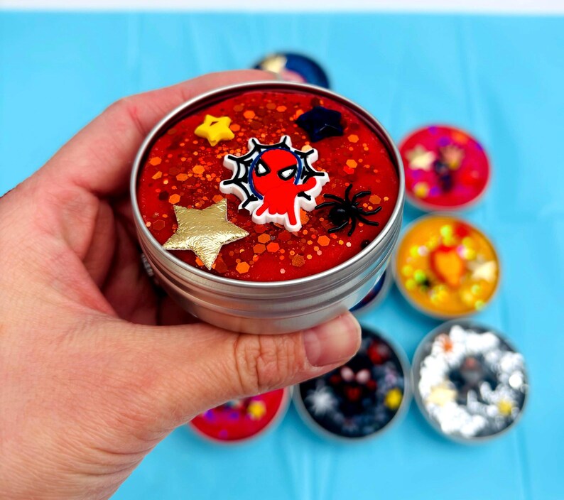 May include: A silver tin filled with red slime, decorated with a web-crawler emblem, gold and yellow stars, and a black spider. Other tins with colourful contents are in the background.