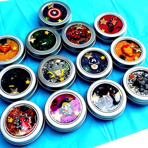 May include: A selection of small, round, silver tins filled with colourful, glittery substances. Each tin features a different superhero character, including American superhero, web crawler, and Metal superhero Man. The tins are arranged on a bright blue surface.