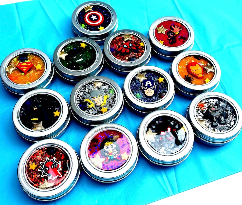 May include: A selection of small, round, silver tins filled with colourful, glittery substances. Each tin features a different superhero character, including American superhero, web crawler, and Metal superhero Man. The tins are arranged on a bright blue surface.