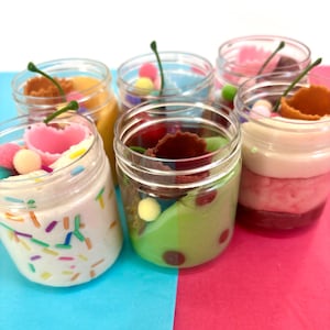 Peut inclure: Une collection de six pots en plastique transparent remplis de slime coloré, chacun surmonté d'un petit morceau de cône gaufré marron, d'une tige verte et d'éléments décoratifs variés comme des vermicelles, des pompons et des cœurs.