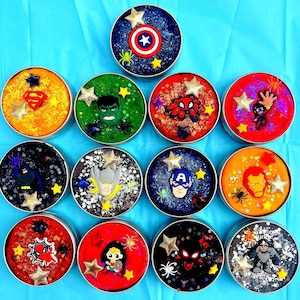 Twelve round metal tins filled with colourful, glittery homemade playdough . Each container features a different superhero emblem or character, including all superhero’s, American hero, and Metal Man, with star and spider accents.
