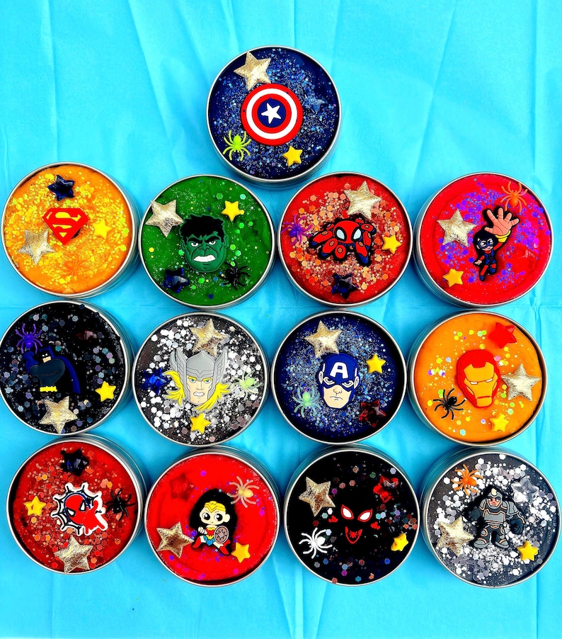 Twelve round metal tins filled with colourful, glittery homemade playdough . Each container features a different superhero emblem or character, including all superhero’s, American hero, and Metal Man, with star and spider accents.