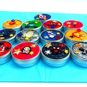 Puede incluir: Una colecci&oacute;n de peque&ntilde;os botes redondos plateados llenos de slime colorido. Cada bote est&aacute; decorado con adornos tem&aacute;ticos de superh&eacute;roes, incluyendo estrellas, ara&ntilde;as y figuras de personajes. Los colores del slime var&iacute;an.