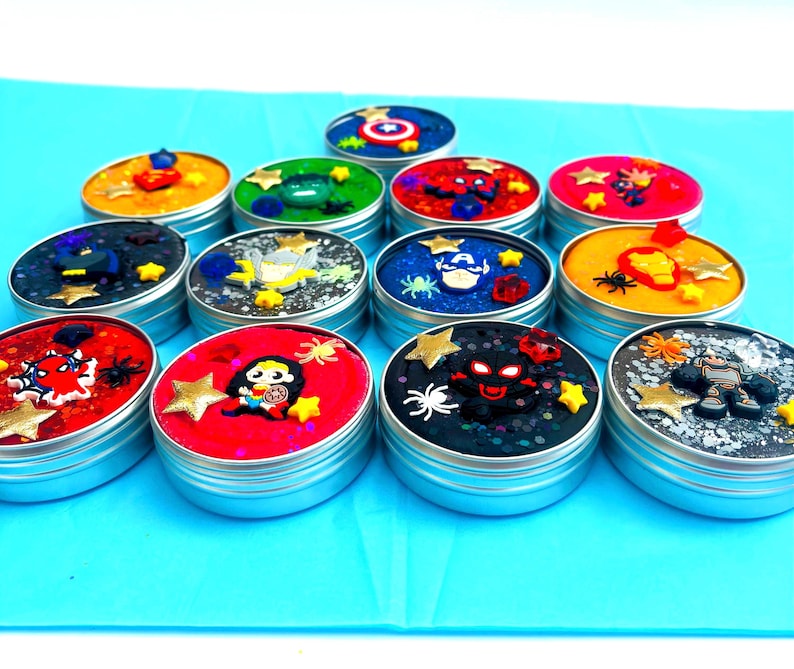 Puede incluir: Una colecci&oacute;n de peque&ntilde;os botes redondos plateados llenos de slime colorido. Cada bote est&aacute; decorado con adornos tem&aacute;ticos de superh&eacute;roes, incluyendo estrellas, ara&ntilde;as y figuras de personajes. Los colores del slime var&iacute;an.