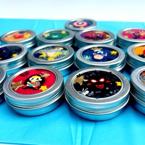 Puede incluir: Una colecci&oacute;n de peque&ntilde;os botes con tapas plateadas llenos de sustancias coloridas y brillantes. Cada bote presenta un dise&ntilde;o diferente con tem&aacute;tica de superh&eacute;roes. Los botes est&aacute;n dispuestos sobre una superficie azul brillante.